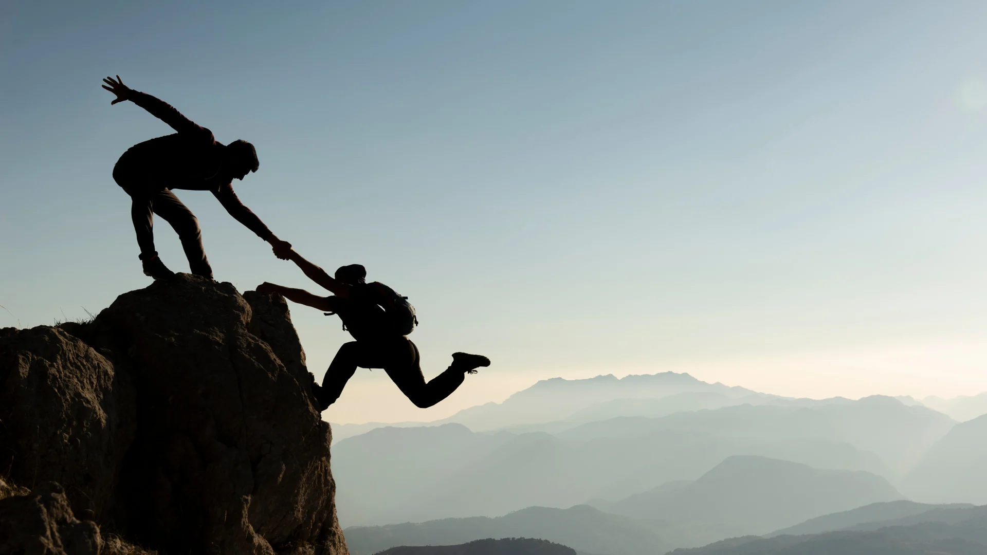 A person on top of a mountain helps a second person make the climb