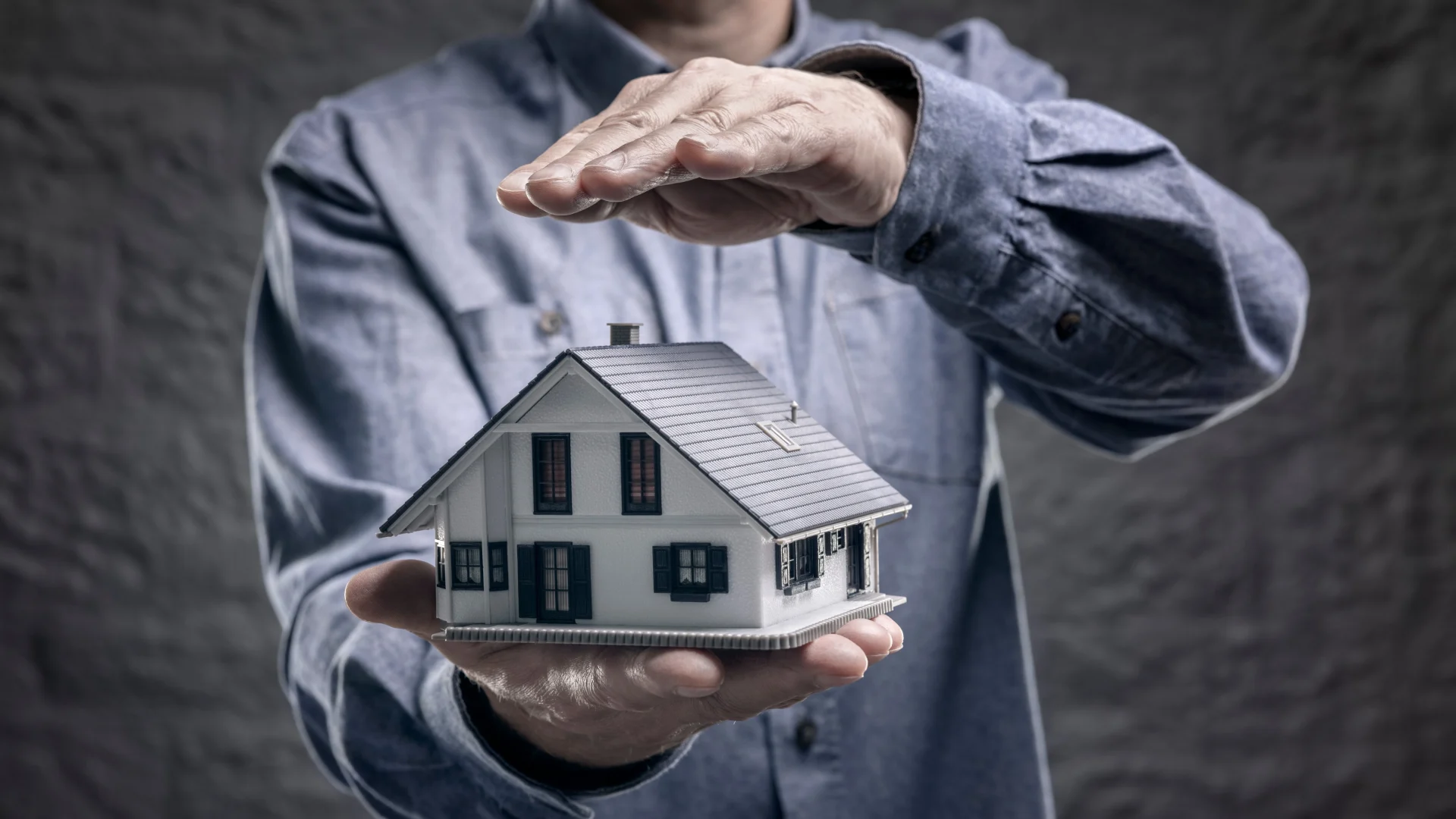 Image of a man holding a model house in one hand and shielding it from above with his other hand