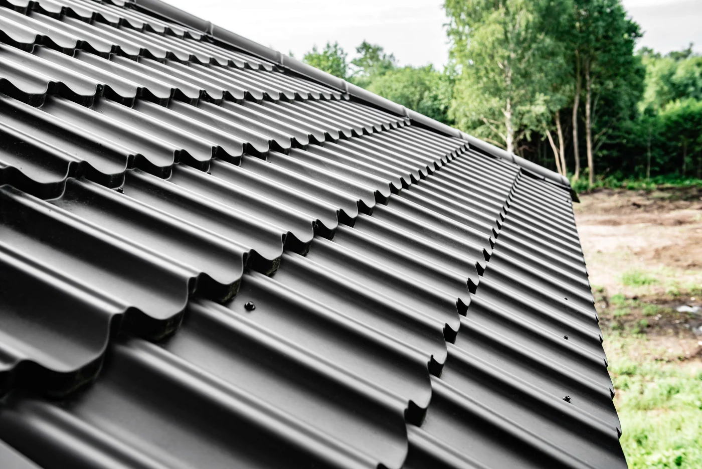 Construction Of The Roof Of The House. Metal Tiles. A close-up of black metal roofing shingles with the woods in the background.