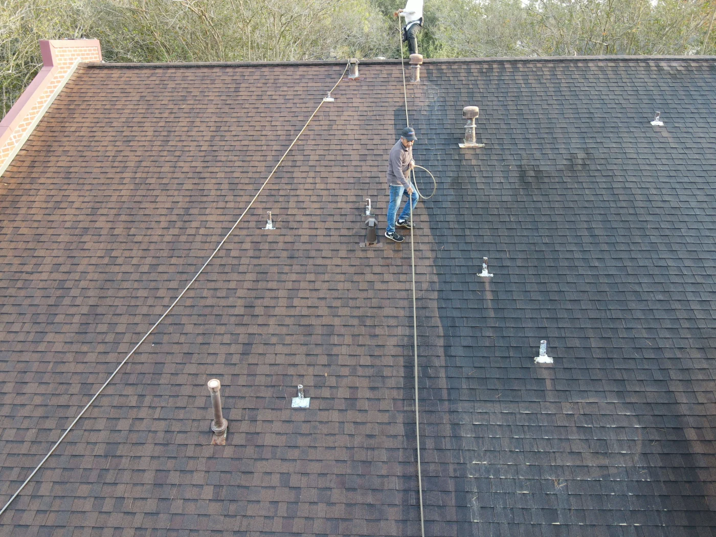Reliable Roof Cleaning | Clear Lake City, TX | Brinkmann Quality Roofing Services An aerial view demonstrates the striking progress of a professional roof cleaning service, showing the clear line where dark stains and debris are being systematically washed away from old asphalt shingles.