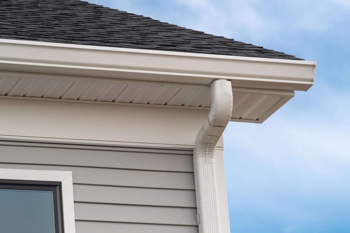 A close-up view of a white gutter and downspout beneath a dark shingle roof on a gray-sided home against a bright blue sky.