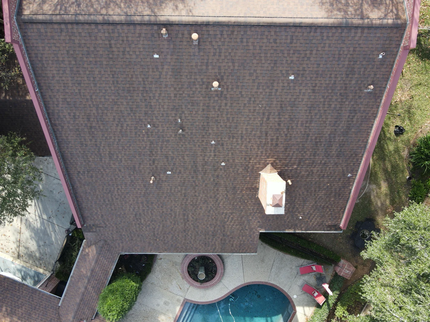 Default Aerial view of a large brown shingle roof with vents, chimney, shaded areas, and a backyard pool beside trees and a patio