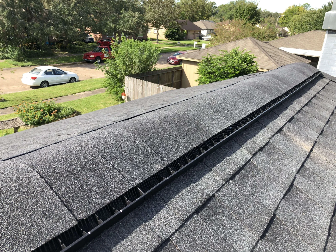Remarkable Roof Maintenance Fort Bend Tx Brinkmann Quality Roofing Close-up of a dark asphalt shingle roof with a ridge vent beside a chimney, overlooking a tree-lined neighborhood.