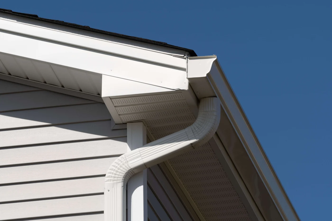 Close-up of white gutters and a curved downspout beneath a shingle roof, with vented soffits, gray siding, and a clear blue sky.