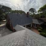 Gray shingle roofs cover a brick home with a chimney, patio, driveway, mature trees, and sunlight breaking through a cloudy sky.