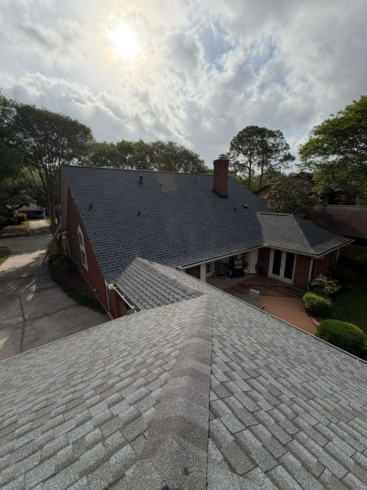 Understanding Roof Insurance Clear Lake City Tx Brinkmann Quality Roofing Gray shingle roofs cover a brick home with a chimney, patio, driveway, mature trees, and sunlight breaking through a cloudy sky.