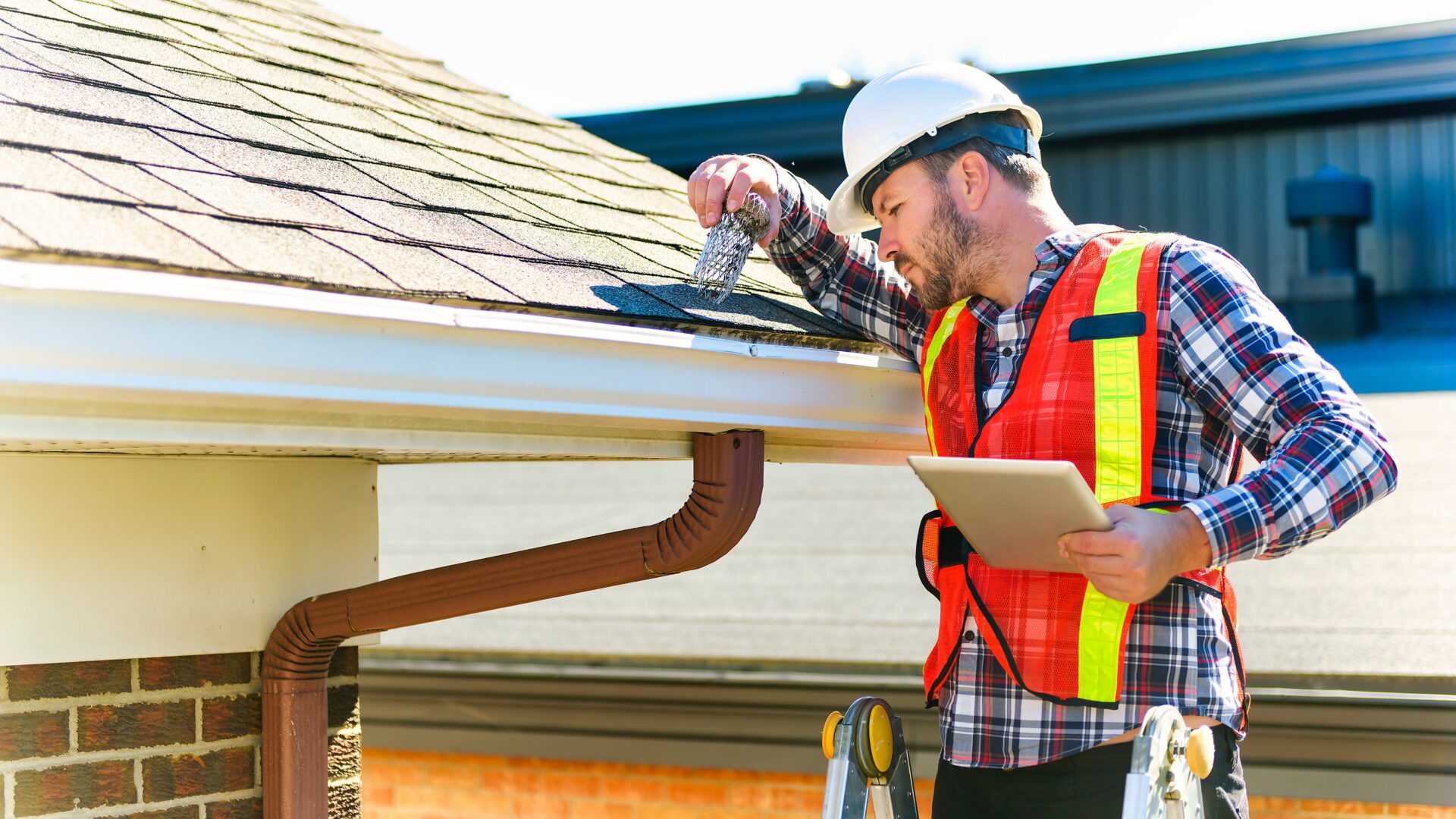 A roofer with a hardhat on inspects the downspout of a gutter system