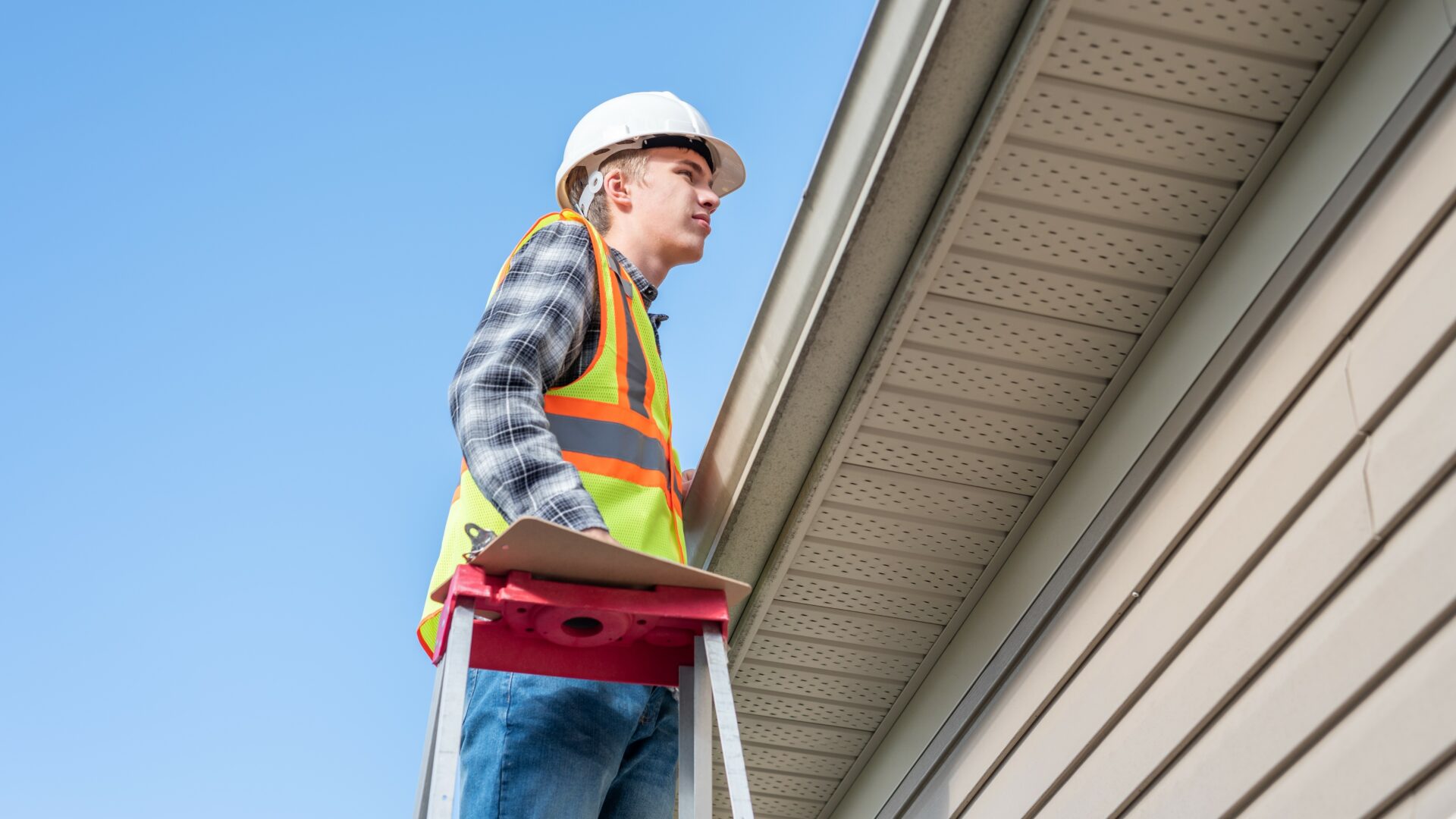 A roofer with a hardhat on inspects the edge of a roof